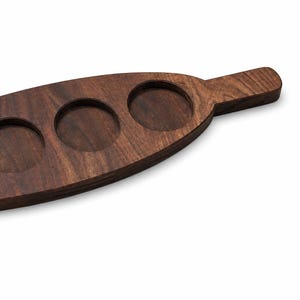 Puede incluir: Una tabla de servir de madera de color marrón oscuro, de forma alargada, con un acabado liso y un mango en un extremo. La tabla presenta cuatro hendiduras circulares dispuestas en fila, diseñadas para sostener objetos pequeños.