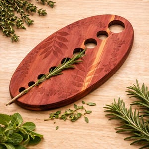 May include: Oval-shaped wooden serving board with leaf pattern and four holes. A sprig of rosemary is placed in the holes. Fresh herbs, including thyme, rosemary, and mint, are arranged around the board on a wooden surface.
