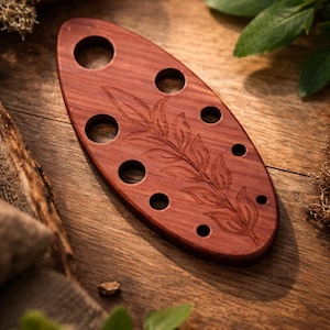 May include: A reddish-brown wooden oval-shaped item with multiple circular holes and a carved leaf design. The item is displayed on a wooden surface, suggesting a decorative or functional object.