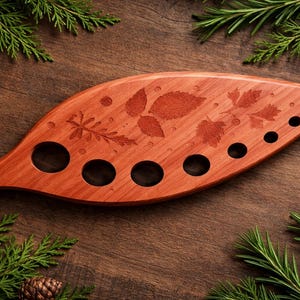 May include: A leaf-shaped wooden serving tray with engraved leaf and vine designs. The tray has several circular cutouts and is surrounded by evergreen branches and pinecones, creating a rustic, natural aesthetic.