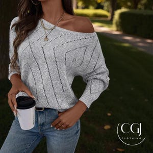 May include: A light grey off-the-shoulder jumper with a geometric pattern. The jumper has long sleeves and is paired with blue jeans. A person is holding a white coffee cup with a black lid. A gold necklace is visible.