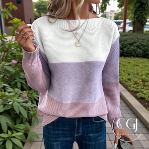 May include: A colour-block sweater in white, lavender, and pink. The long-sleeved knitwear has a boat neck and is paired with blue jeans. The image also shows a person wearing the sweater, rings, and a necklace.