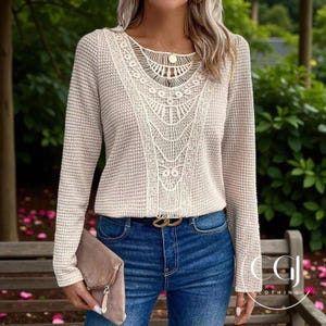 Könnte beinhalten: Beiger Langarmpullover mit cremefarbenem Spitzeneinsatz. Der Pullover hat ein strukturiertes Strickmuster und einen runden Ausschnitt. Das Model trägt eine blaue Jeans und einen goldenen Gürtel.