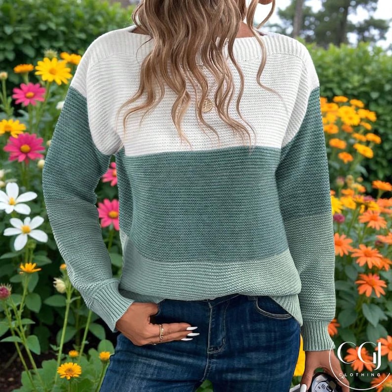 May include: A colour-block sweater in white, green, and light green. The long-sleeved sweater has a round neckline and is paired with blue jeans. The background features colourful flowers and foliage.