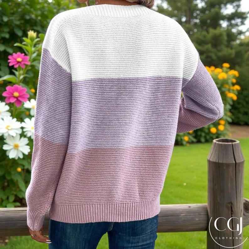 May include: A colour-block sweater in white, lavender, and mauve. The sweater has a ribbed knit texture and long sleeves. The back view shows the sweater's design, with the colour blocks horizontally across the chest and sleeves.