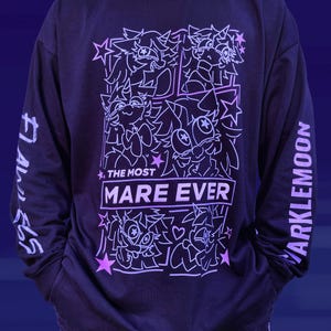 May include: A dark purple long-sleeve shirt featuring a light purple graphic print on the front with multiple cartoon pony illustrations, stars, and the text THE MOST MARE EVER. The left sleeve displays the text FLAWLESS and the right sleeve says MARKLEMOON.
