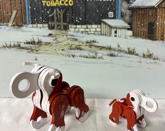 May include: Two chain link sculptures of a dog, one large and one small. The sculptures are painted red and white. The background is a winter scene with a barn that says "MAIL POUCH TOBACCO".
