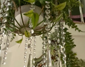 Puede incluir: Una maceta colgante con una estructura de alambre esférica que contiene una maceta blanca con follaje verde. La estructura está adornada con múltiples hilos de cuentas de cristal facetadas transparentes y largos colgantes de vidrio en forma de lágrima.