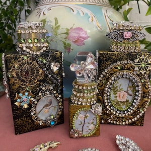 May include: Three ornate, embellished perfume bottles with jewel-encrusted tops and decorative frames. The bottles feature intricate designs, including a bird, floral, and butterfly motifs. Additional jeweled brooches are displayed in the foreground.