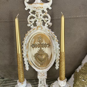 May include: White and gold ornate wall sconce with two candle holders and a decorative oval panel featuring a gold heart and cross design.