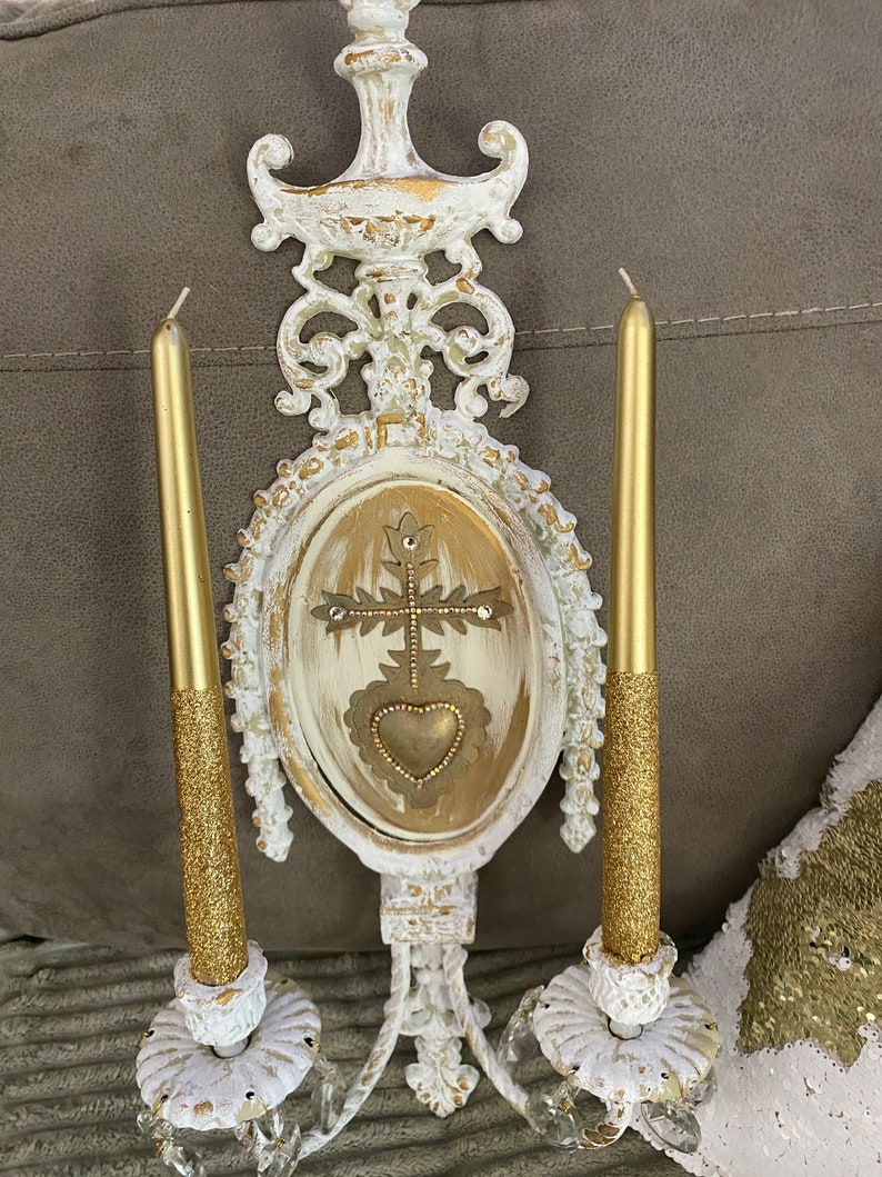 May include: White and gold ornate wall sconce with two candle holders and a decorative oval panel featuring a gold heart and cross design.