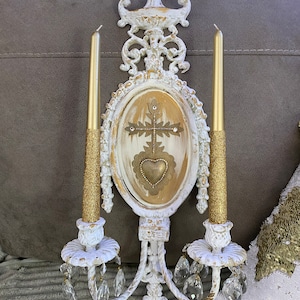 May include: White and gold ornate wall sconce with two candle holders and crystal drops. The sconce features a central oval panel with a gold cross and heart design.
