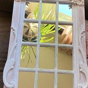 May include: A rectangular mirror with a window pane design. The frame is painted in a distressed white and pink finish, with decorative floral and scroll accents. The mirror reflects a view of greenery and a curtain.