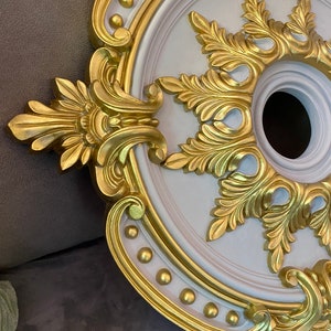 Op de afbeelding: Osiers plafondrozet met een wit midden en goudkleurige rand. Het ontwerp heeft gedetailleerde bloemen- en krulpatronen. De rozet heeft een cirkelvorm met een centrale opening voor een armatuur.