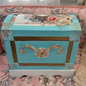 May include: A rectangular turquoise wooden chest with a curved lid, featuring gold-painted trim, ornate floral relief carvings, and a central decorative medallion. The lid displays a printed floral design with pink roses and a cameo-style oval frame.