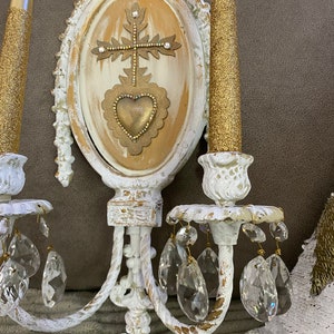 May include: White wall sconce with gold accents and crystal drops. The sconce features a central oval panel with a gold heart and cross design.