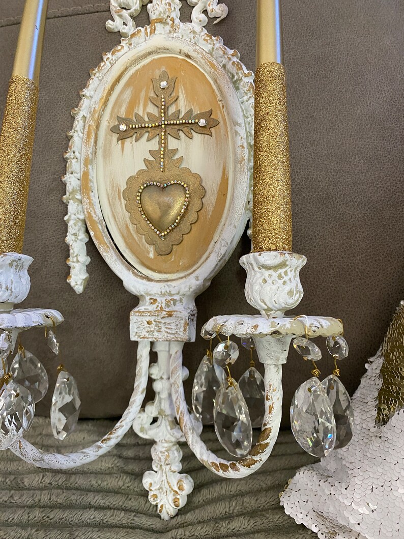 May include: White wall sconce with gold accents and crystal drops. The sconce features a central oval panel with a gold heart and cross design.