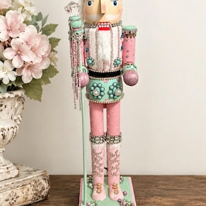 May include: A decorative nutcracker figure featuring a pink faux fur hat, a light blue jacket with turquoise floral accents, and pink trousers. The figure holds a staff topped with a clear crystal and is adorned with rhinestones and pearls.