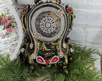 Könnte beinhalten: Verzierte dekorative Uhr in Schwarz und Gold mit einem kreisförmigen Zifferblatt, das mit Strasssteinen und roten Akzenten verziert ist. Die Uhr hat ein detailliertes Design mit einem schneeflockenartigen Schmuck in der Mitte. Die Uhr steht auf grünem Laub.