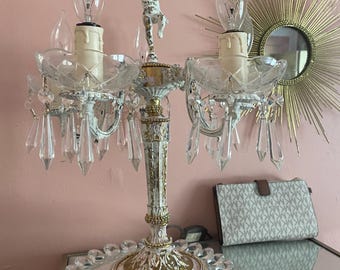 May include: A vintage white and gold candelabra with three candle holders, clear crystal accents, and a small figurine. The candelabra sits on a mirrored surface with a decorative glass tray.