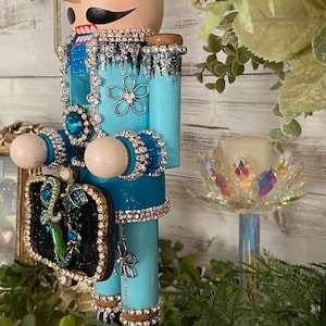 May include: A decorative wooden nutcracker in a light blue uniform, adorned with rhinestones and jewels. The nutcracker has a tan face, a brown hat, and black boots. It holds a small, ornate black box with jewels. A candle holder and flowers are in the background.
