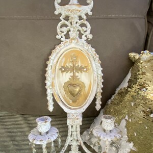 May include: White metal wall sconce with two candle holders and a decorative oval center piece with a gold cross and heart. The sconce has crystal accents.