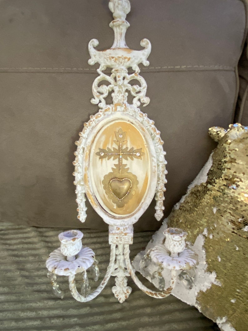May include: White metal wall sconce with two candle holders and a decorative oval center piece with a gold cross and heart. The sconce has crystal accents.