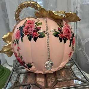 May include: A decorative light pink pumpkin featuring a floral rose pattern, accented with gold-painted leaves and a gold stem. The pumpkin is embellished with vertical rows of clear rhinestones and a dangling crystal charm at the center front.