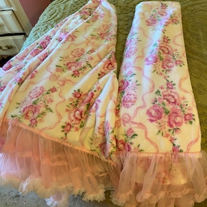 May include: A cream-colored blanket with a pink floral pattern and pink ribbon accents. The blanket has a ruffled pink tulle trim. The blanket is folded and displayed on a green surface.