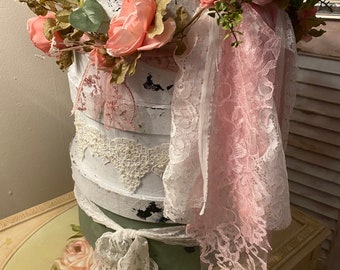 May include: A stack of three round, white, distressed boxes with a pink and white lace trim. The boxes are decorated with a pink floral garland and a pink ribbon.