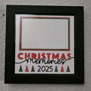 May include: A black framed print with the words "CHRISTMAS memories 2025" in red and black script. The print has a white background with a red and green border and green Christmas tree graphics.
