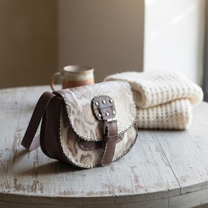 Peut inclure: Un sac bandoulière en cuir marron et à motif peau de serpent avec une fermeture à boucle. Le sac a une sangle marron et est posé sur une table en bois vieilli. Une tasse et une couverture crème pliée sont en arrière-plan.