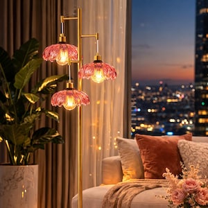 May include: A gold floor lamp with three pink glass shades, illuminating a living room. The lamp stands tall, casting a warm glow on the surrounding decor, including a sofa, rug, and side table. The background features a cityscape at dusk.