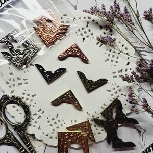 Könnte beinhalten: Eine Sammlung dekorativer Metallecken in verschiedenen Ausführungen, darunter Bronze, Silber und Schwarz. Die Ecken sind auf einer weißen Spitze angeordnet, zusammen mit einer Vintage-Schere und Trockenblumen. Das Bild enthält auch ein Etikett mit dem Text "Eckenschutz".