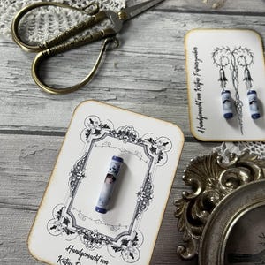 Könnte beinhalten: Eine antike Schere mit verzierten Griffen liegt neben zwei dekorativen Karten. Eine Karte zeigt ein gerahmtes Bild eines kleinen, zylindrischen Objekts mit einem Porträt. Die andere Karte zeigt ein Design mit hängenden Ornamenten.