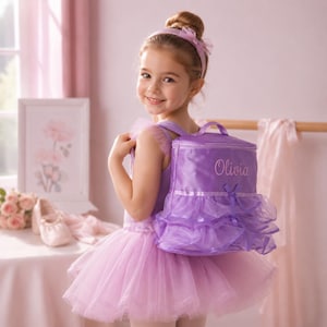 Puede incluir: Una niña con tutú y diadema rosas sonríe, llevando una mochila morada con el nombre "Olivia" bordado. La mochila tiene un diseño de falda con volantes. El fondo es de un rosa suave.