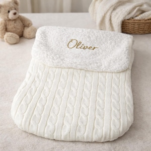 May include: Cream-coloured baby sleep sack with a cable knit design. The top section is a soft, plush material, embroidered with the name "Oliver" in gold thread. A teddy bear is visible in the background.