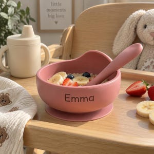 Può includere: Una ciotola a ventosa in silicone rosa polvere con il nome Emma stampato sul davanti, contenente farina d'avena guarnita con fette di banana, mirtilli e fragole, accompagnata da un cucchiaio in silicone abbinato e un bicchiere beige.