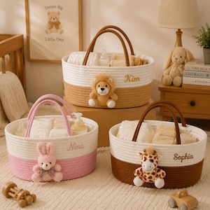 Puede incluir: Tres cestas de almacenamiento de cuerda trenzada con asas. Una cesta es blanca y marrón con un peluche de león, otra es blanca y rosa con un peluche de conejo, y la tercera es blanca y marrón con un peluche de jirafa. Cada cesta tiene un nombre bordado.