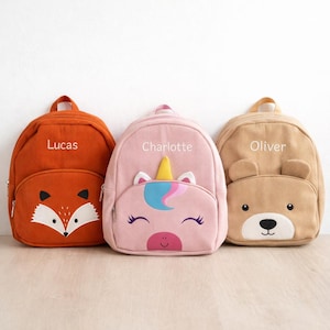 May include: Three corduroy backpacks in orange, pink, and tan. The orange backpack has a fox face, the pink one a unicorn, and the tan one a bear face. Each backpack has a name embroidered on it: Lucas, Charlotte, and Oliver.