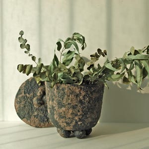 May include: A textured, dark brown and tan ceramic pot with a lid, filled with green and brown eucalyptus branches. The pot has a rounded shape and sits on three small, round feet. The lid is placed to the side.