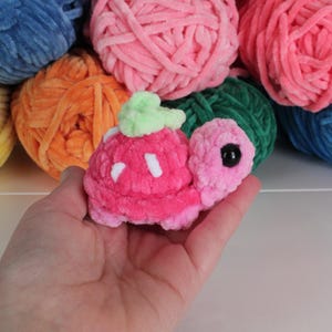 May include: A handmade plush turtle in shades of pink, with a green leaf-like detail on its back. The turtle is held in a hand, with colorful yarn balls in the background. The yarn balls are in shades of blue, orange, pink, green, and yellow.