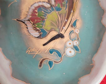May include: Oval decorative dish featuring a hand-painted butterfly design. The butterfly has colourful wings and is set against a teal background with gold accents. The dish has a white border and the signature "Adry".
