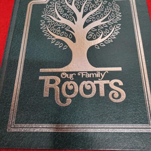 Könnte beinhalten: Ein dunkelgrünes Familienchronikbuch mit den Worten "Our Family Roots" in silbernen Buchstaben. Das Cover zeigt ein silbernes Baumdesign innerhalb eines silbernen Rahmens. Das Buch ist geschlossen und liegt auf einer roten Oberfläche.