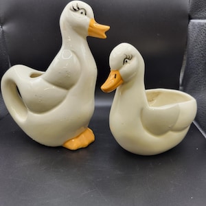 May include: Two ceramic duck figurines. One is a tall pitcher with a handle, and the other is a small bowl. Both are off-white with orange beaks and feet. The ducks have black eye details and are positioned on a dark surface.