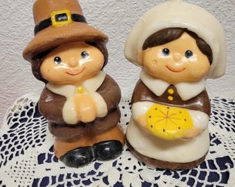 May include: Two vintage Thanksgiving candles. One candle is a pilgrim man wearing a brown hat with a black band and buckle. The other candle is a pilgrim woman wearing a white bonnet and holding a yellow pie. Both candles have painted faces.