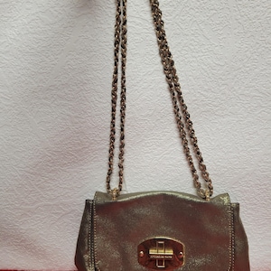 Può includere: Una borsa a tracolla color oro metallizzato con una tracolla a catena. La borsa ha una forma rettangolare con chiusura a patta e una chiusura dorata. La tracolla a catena è anch'essa dorata. La borsa è su uno sfondo bianco strutturato.