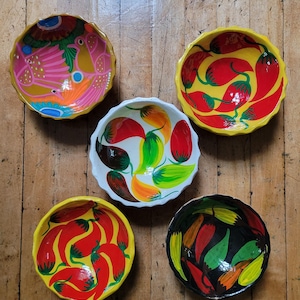 Könnte beinhalten: Fünf handbemalte Keramikschalen mit gewellten Rändern. Die Schalen weisen lebendige Designs von Vögeln, Chilischoten und abstrakten Mustern in einer Vielzahl von Farben auf, darunter Rot, Gelb, Rosa, Grün und Schwarz. Die Schalen sind auf einer Holzoberfläche angeordnet.