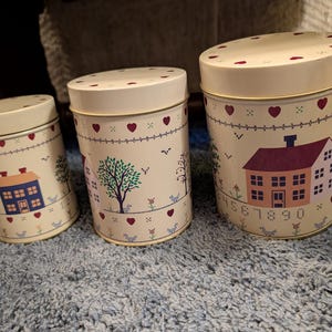 May include: Three cream-colored metal canisters with lids. Each canister features a house and tree design with hearts and numbers. The canisters are different sizes and sit on a blue surface.