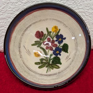 May include: A round ceramic plate with a floral design. The plate features a cream-colored center with a bouquet of flowers in shades of red, yellow, and blue. The rim has a blue and brown border.
